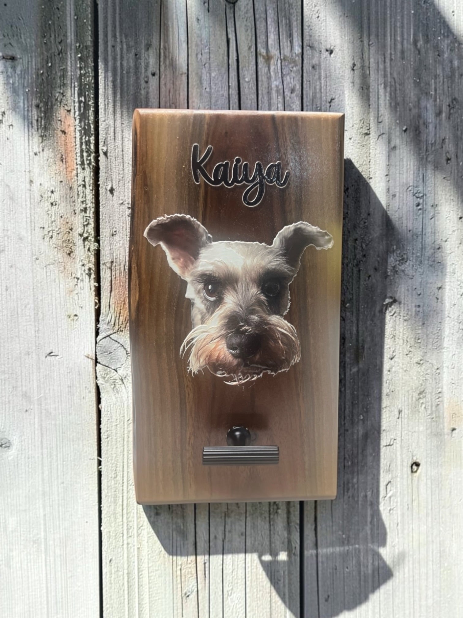 A rectangular wooden plaque mounted on weathered gray wood siding, featuring a printed or carved portrait of a small dog with the name 'Kaiya' above it and a small metal slot at the bottom.