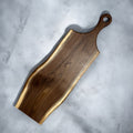 A rustic live-edge wooden serving board with a dark brown grain and a light natural wood border, shown diagonally on a textured gray concrete surface.