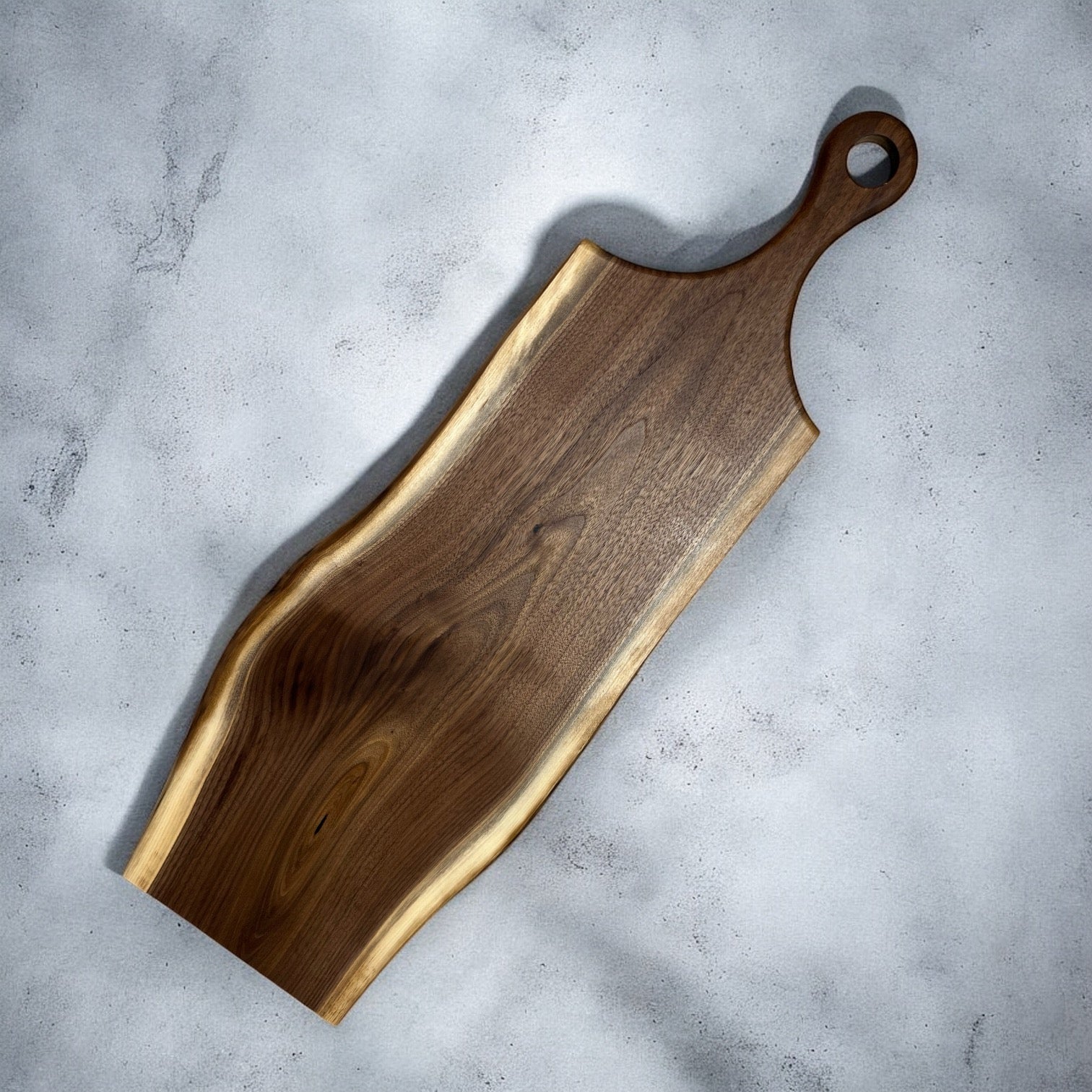 A rustic live-edge wooden serving board with a dark brown grain and a light natural wood border, shown diagonally on a textured gray concrete surface.