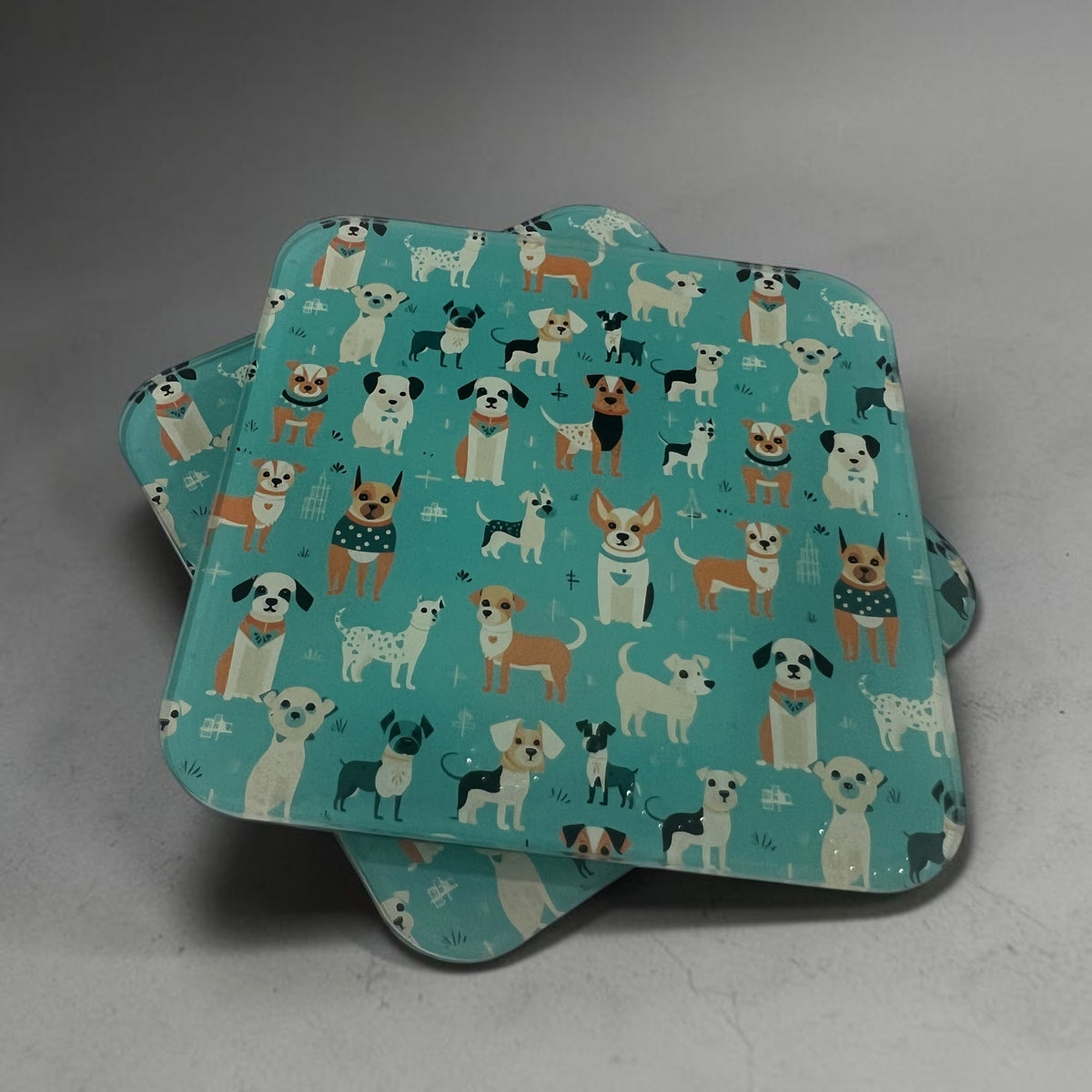A stack of square coasters with rounded corners featuring a repeating pattern of various dog illustrations on a teal background, set against a neutral gray studio backdrop.