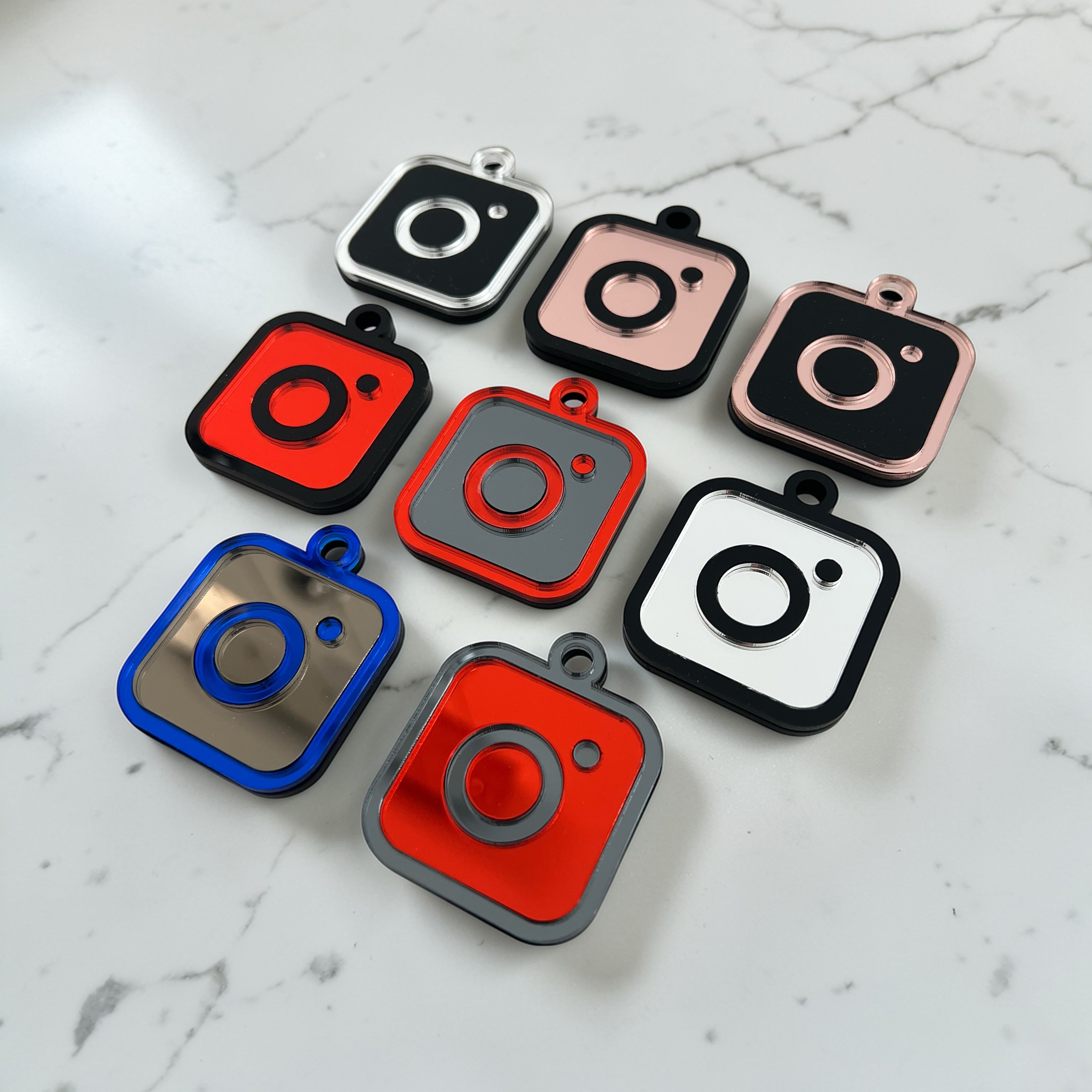 Eight Instagram social media tags arranged on a white marble surface. Each charm has a rounded square body with a small loop at the top to put on a keychain.  Shown in various color combinations including red, black, silver, rose gold, blue and grey.