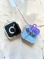 Both sides of an acrylic social media tag is shown, on a white marble surface. One side of the tag shows the Instagram logo that is black with a silver border and the other side is a logo with a purple gradient flower with the text 'Bring Out Your AWESOME!' 