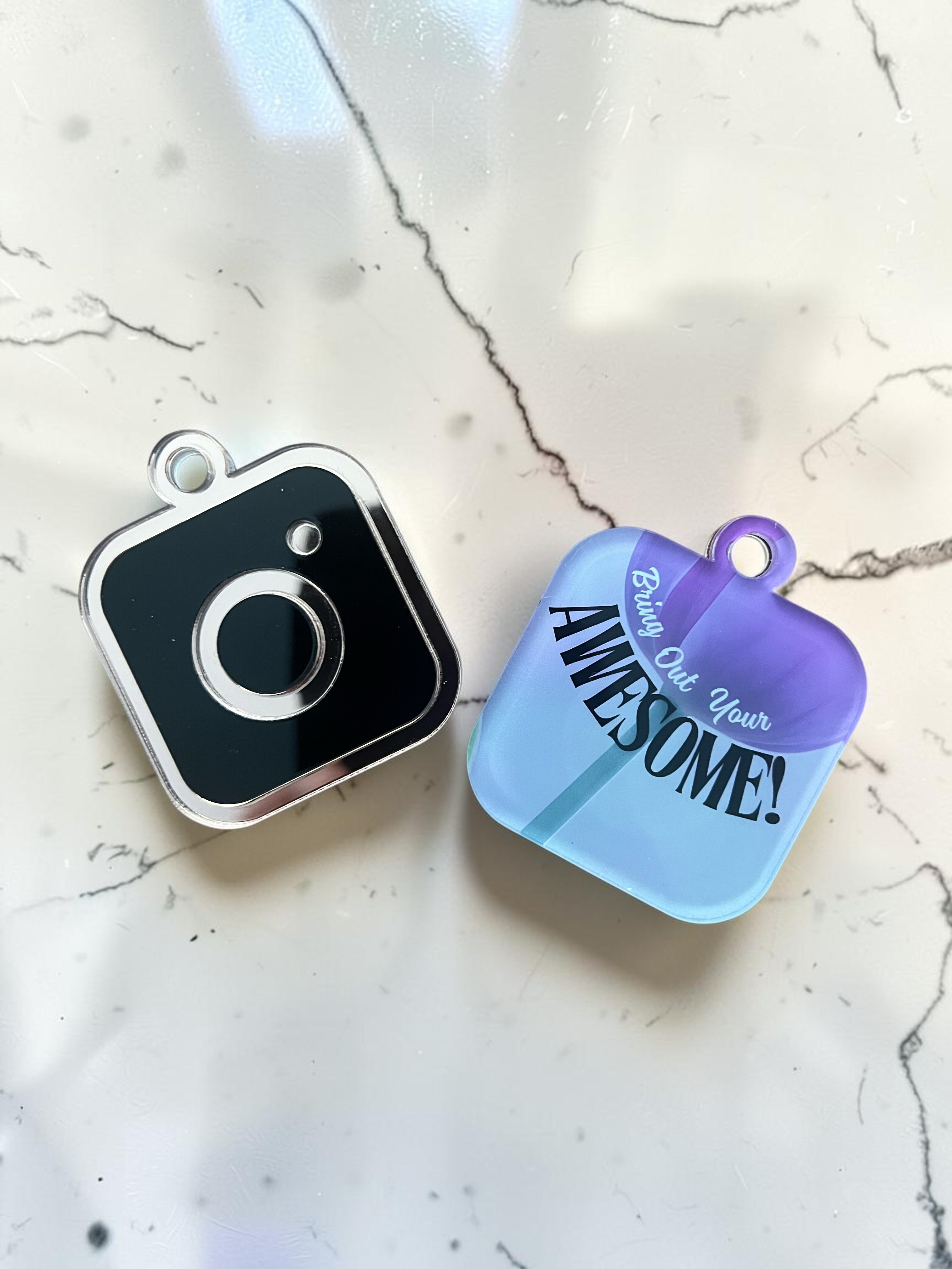 Both sides of an acrylic social media tag is shown, on a white marble surface. One side of the tag shows the Instagram logo that is black with a silver border and the other side is a logo with a purple gradient flower with the text 'Bring Out Your AWESOME!' 