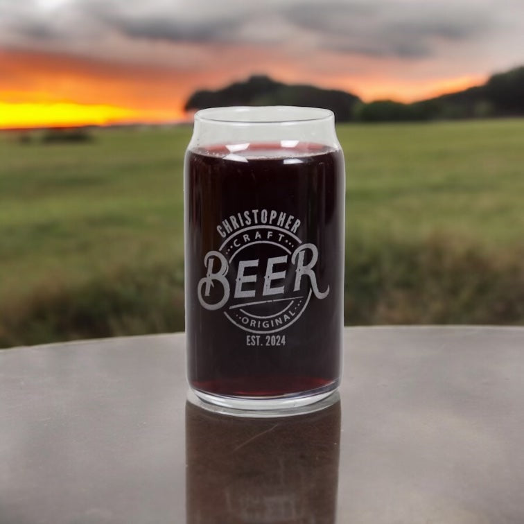 A clear glass can-shaped beer standing on a reflective tabletop with a blurred outdoor field and sunset in the background. The glass has white printed text reading 'CHRISTOPHER CRAFT BEER ORIGINAL EST. 2024