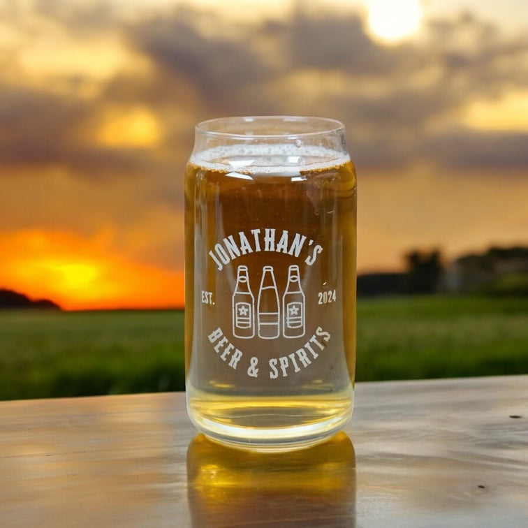 A clear glass can-shaped beer sits on a wooden table outdoors at sunset, with a blurred field and dramatic sky in the background. The glass is printed with the text 'JONATHAN'S BEER & SPIRITS' and three beer bottle illustrations.