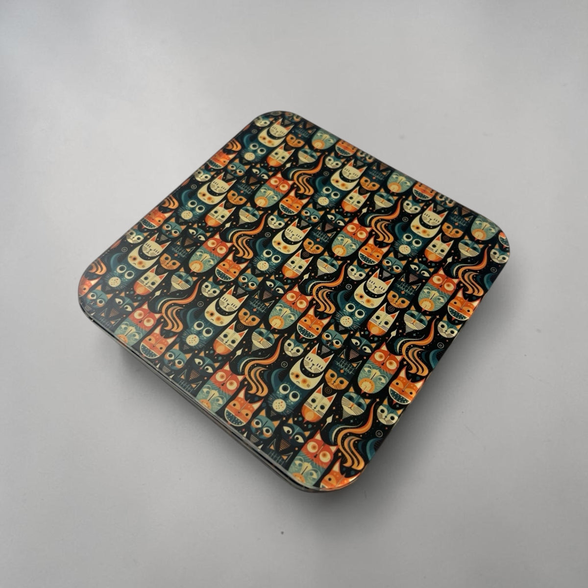 A coaster with rounded corners, featuring a repeating pattern of cute cartoon cats in orange, yellow, and teal on a neutral light gray surface.