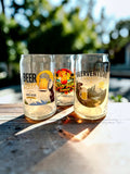 Three clear pint glasses with colorful beer-themed printed designs sit on a wooden table outdoors in bright sunlight, with blurred green foliage in the background.