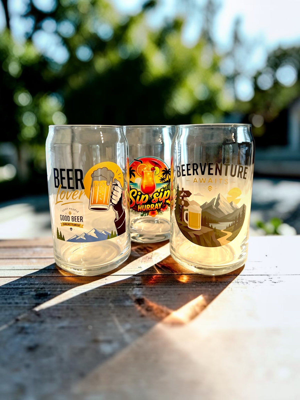 Three clear pint glasses with colorful beer-themed printed designs sit on a wooden table outdoors in bright sunlight, with blurred green foliage in the background.