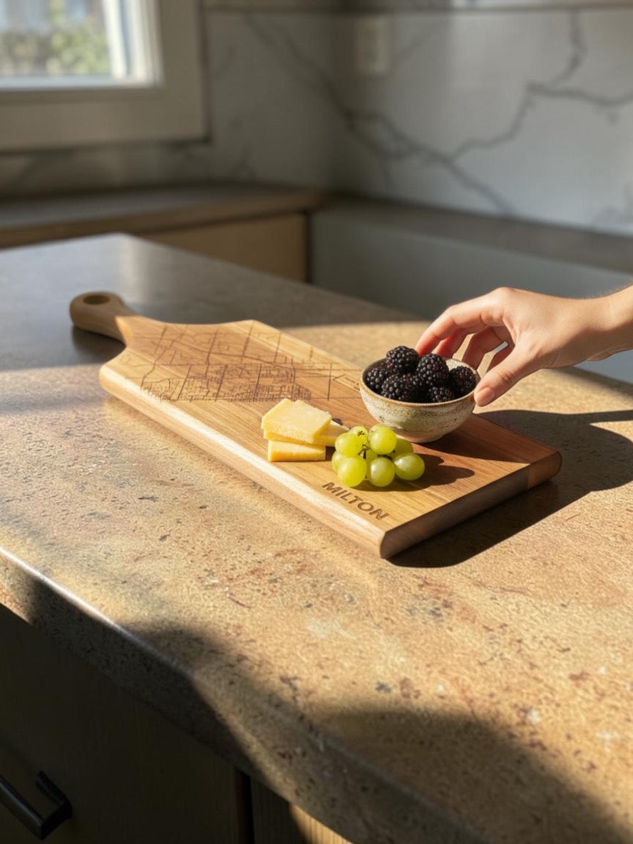 A wooden serving board with a handle and engraved brand name, placed on a kitchen countertop. A hand is placing a bowl of blackberries on top, with cheese and green grapes also on the board.