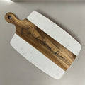 A personalized serving board with rounded corners, featuring a white marble-patterned body and a natural wood handle and center stripe. The wood is engraved with a circular monogram and the name 'katelynroberts brendan'.