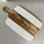 A personalized serving board with rounded corners, featuring a white marble-patterned body and a natural wood handle and center stripe. The wood is engraved with a circular monogram and the name 'katelynroberts brendan'.