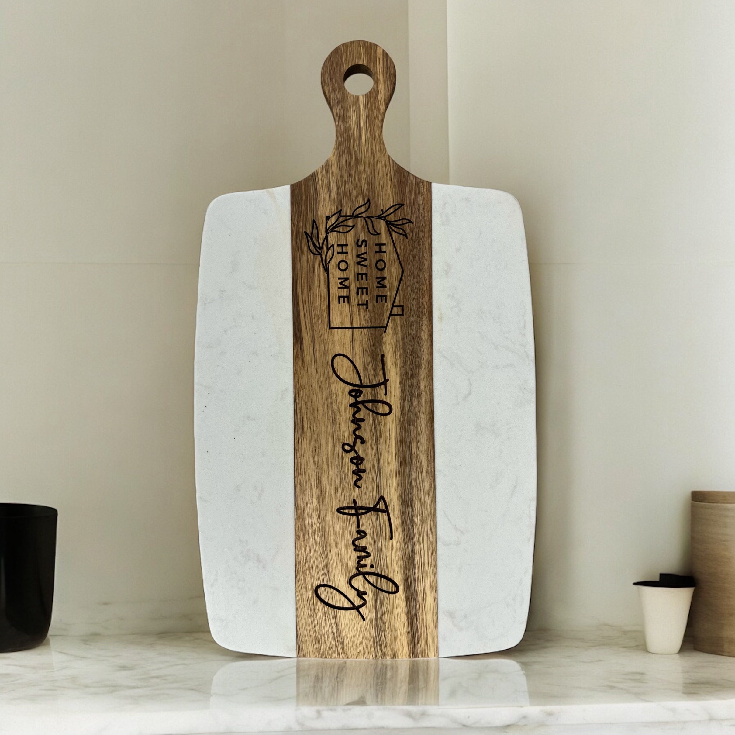 A personalized serving board with rounded corners, featuring a white marble-patterned body and a natural wood handle and center stripe.standing on a marble countertop against a beige tiled wall. The center wood handle features an engraved logo for 'Home Sweet Home' and the name 'Johnson Family' in script.