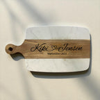 A personalized serving board with rounded corners, featuring a white marble-patterned body and a natural wood handle and center stripe. The wood is engraved with a circular monogram and the name 'Kiki with and attached heart Jensen, September 2025'.