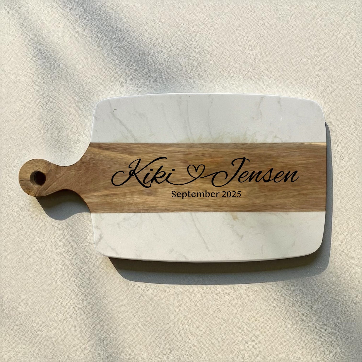 A personalized serving board with rounded corners, featuring a white marble-patterned body and a natural wood handle and center stripe. The wood is engraved with a circular monogram and the name 'Kiki with and attached heart Jensen, September 2025'.