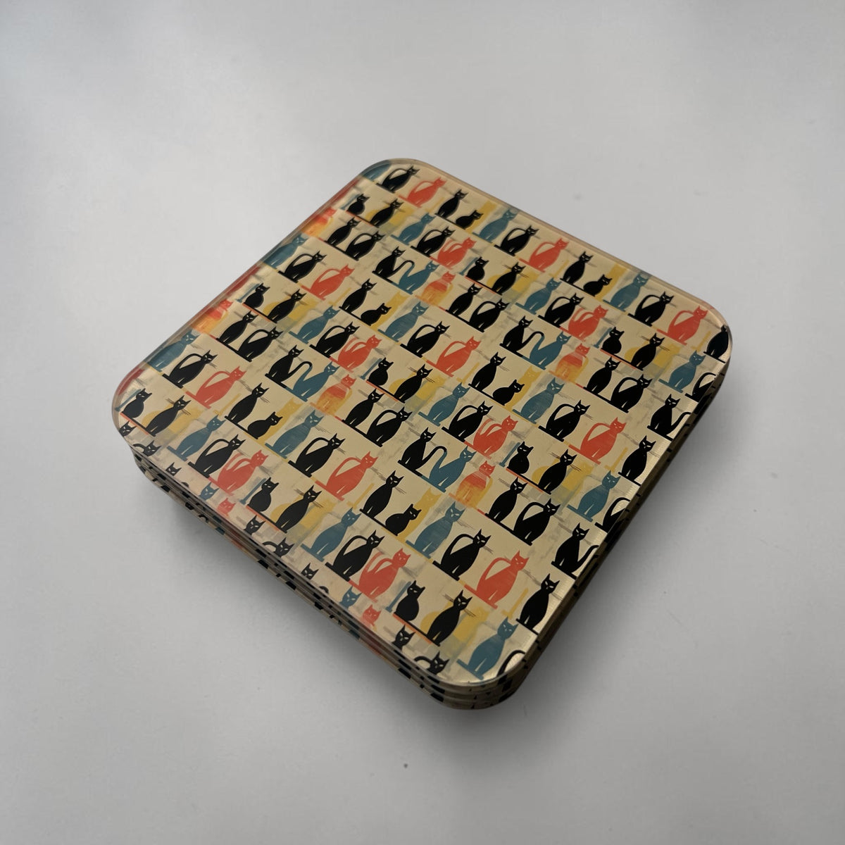 A square coaster with rounded corners, featuring a repeating pattern of stylized cat silhouettes in black, blue, orange, and red on a cream background on a neutral light gray surface.