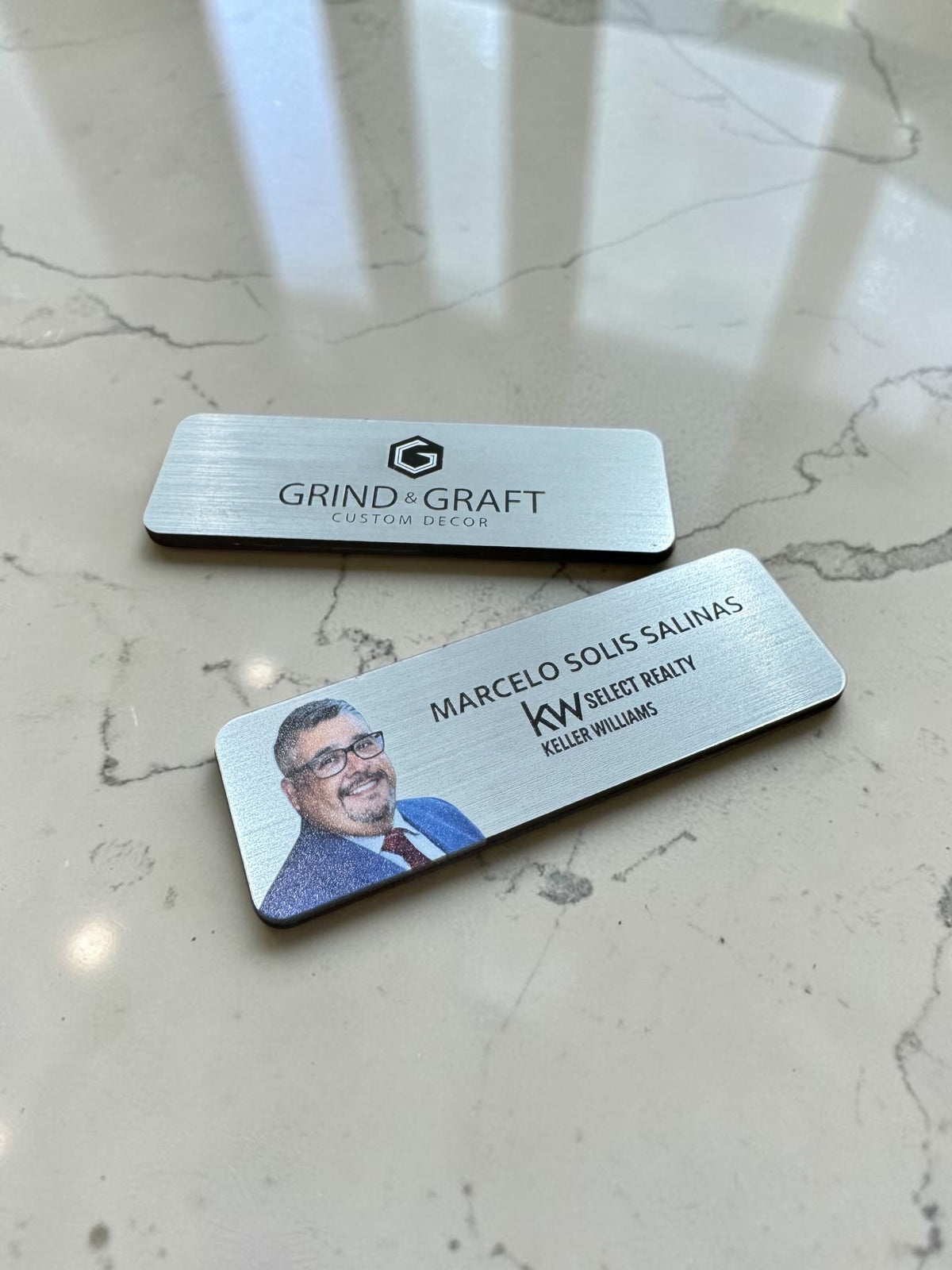 Two custom acrylicl nametags on a light marble surface. The top plate reads 'GRIND & GRAFT CUSTOM DECOR' with a geometric logo, and the bottom plate shows a photo of Marcelo Solis Salinas with 'KW SELECT REALTY' and 'KELLER WILLIAMS'.