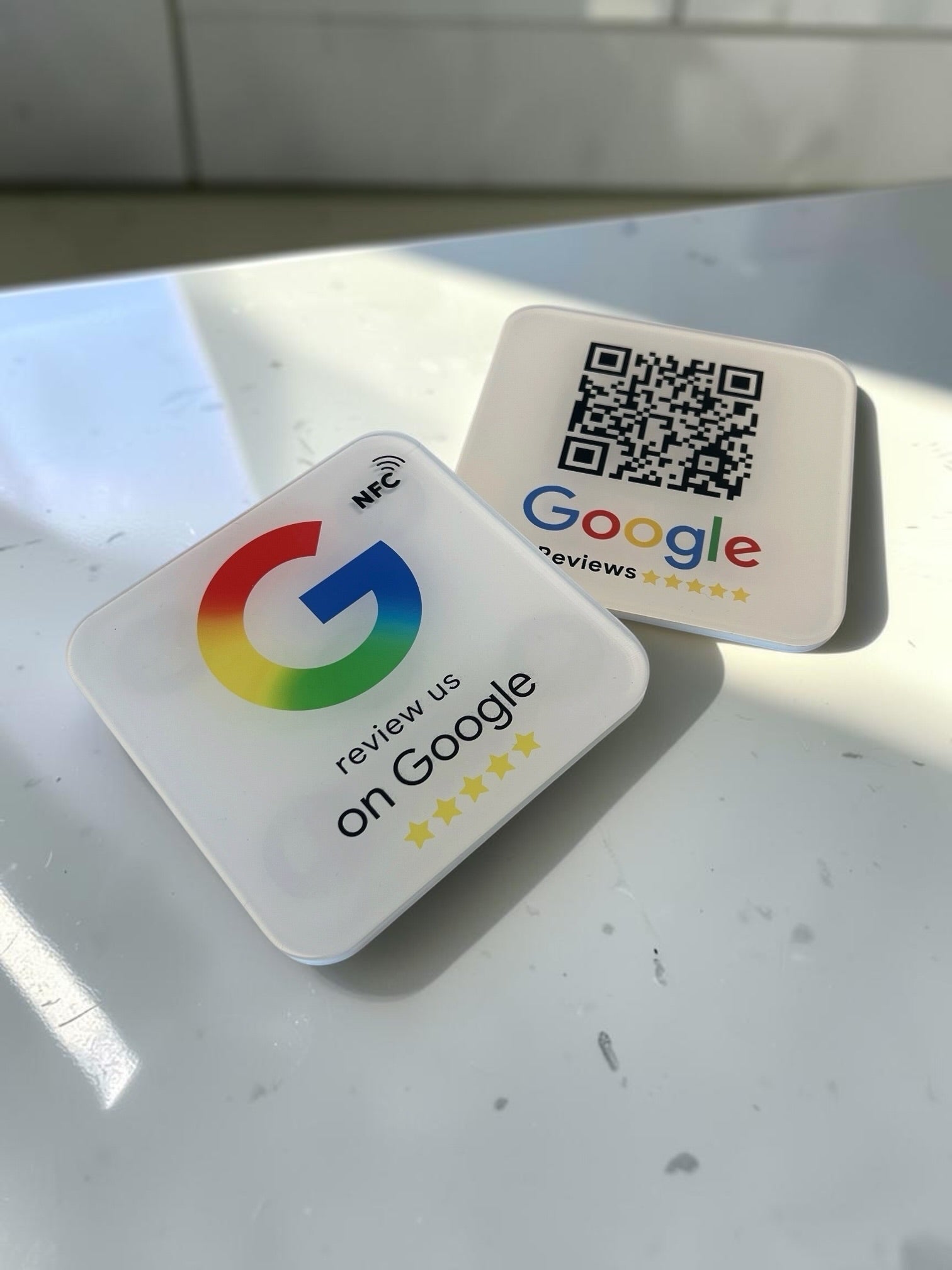Two square white Google review placeholders on a light gray surface. One shows the colorful Google G logo with the text 'review us on Google' and five stars with NFC icon on the top right hand side, and the other shows a QR code above the Google reviews logo.