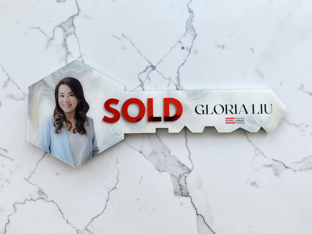 A key-shaped sign on a white marble background, featuring a photo of a woman in a light blue blazer on the key blade, with the word 'SOLD' in bold red letters and the name 'GLORIA LIU' printed in black.