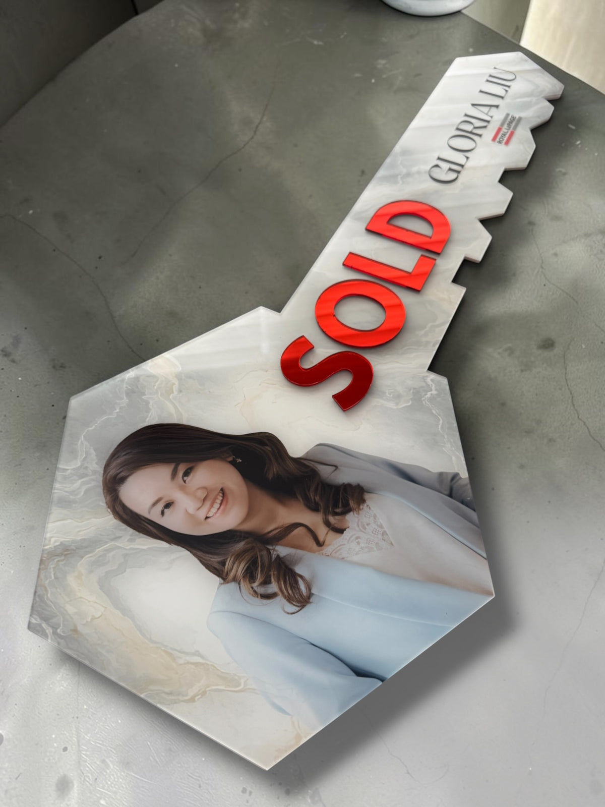 A key-shaped sign on a white marble background, featuring a photo of a woman in a light blue blazer on the key blade, with the word 'SOLD' in bold red letters and the name 'GLORIA LIU' printed in black.
