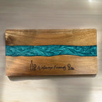 A rectangular wooden serving board with a natural wood grain finish and a horizontal turquoise resin inlay. The board is engraved with the words 'Welcome Friends' in script, flanked by a wine bottle and wine glass icons.