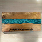 A rectangular wooden serving board with a natural wood grain finish and a horizontal turquoise resin inlay. The board is engraved with the words 'The Kitchen is the Heart of the Home' in script.