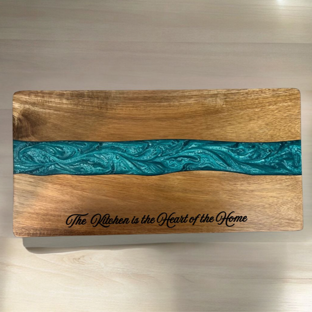 A rectangular wooden serving board with a natural wood grain finish and a horizontal turquoise resin inlay. The board is engraved with the words 'The Kitchen is the Heart of the Home' in script.