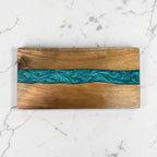 A rectangular wooden serving board with a natural wood grain finish and a central turquoise blue resin river inlay, displayed on a white marble surface.