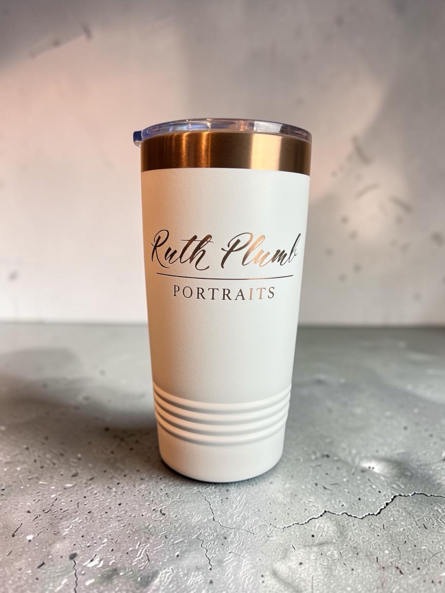 A white insulated travel tumbler with a clear lid and metallic rose gold band, featuring the text 'Ruth Plum Portraits' printed on the front, set on a gray concrete surface.